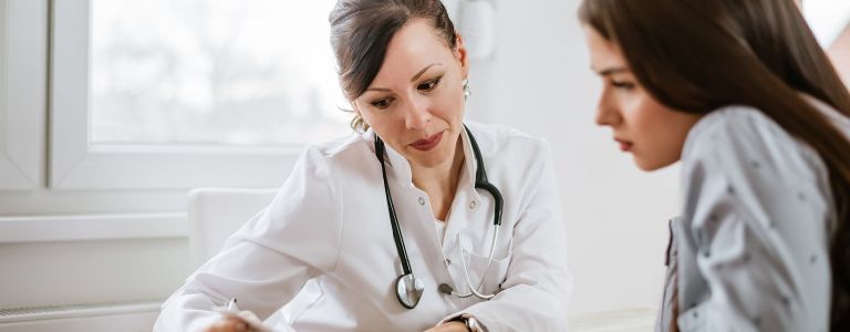 Junge Frau sitzt neben Ärztin, die ihr etwas auf einem Blatt Papier zeigt